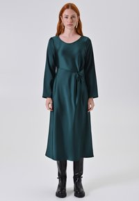 Lungo abito in satin verde con maniche lunghe, scollatura a barca e dettaglio a nodo in vita. Indossato con stivali neri fino al ginocchio.