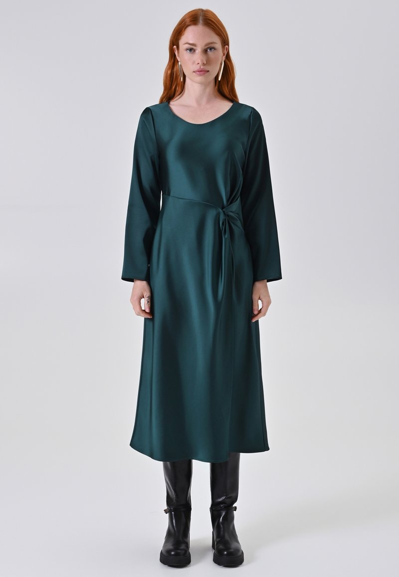 Lungo abito in satin verde con maniche lunghe, scollatura a barca e dettaglio a nodo in vita. Indossato con stivali neri fino al ginocchio.
