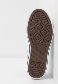 Sula av en vit och blå Converse All Star-sneaker som visar ett brunt mönstrat mönster med logotyp, placerad vertikalt mot en vit yta.