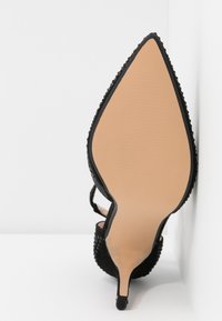 Gros plan sur la semelle d'une chaussure à talon haut noire à bout pointu avec des côtés texturés, posée sur un fond blanc.