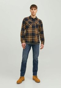 Camisa de cuadros marrón y azul marino con dos bolsillos en el pecho, combinada con jeans azules y botas amarillas. Modelo de pie contra un fondo sencillo.