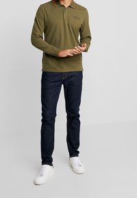 Oliwkowa, długorękawowa koszulka polo z pomarańczowo-czerwonymi paskami na kołnierzyku, w połączeniu z ciemnoniebieskimi dżinsami i białymi trampkami.