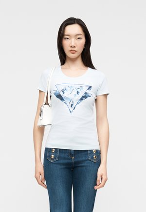 Mujer con una camiseta blanca de manga corta con un gráfico de triángulo azul, jeans azules con botones dorados y un bolso blanco de hombro.