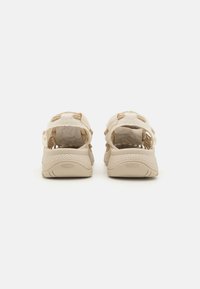 Sandales de sport beiges avec une tige texturée, un talon ouvert et des accents beiges contrastants. Présente une semelle perforée pour la respirabilité.