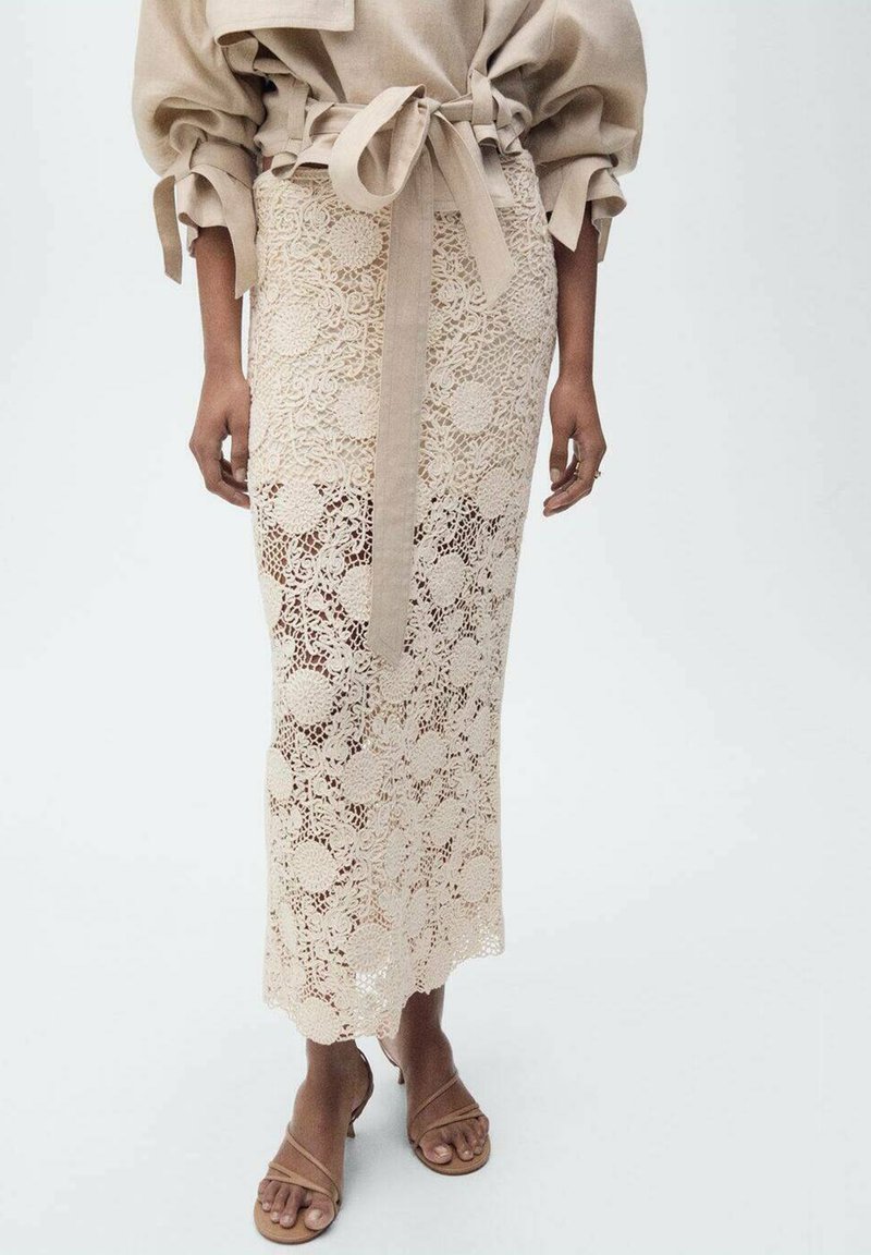 Gonna midi in pizzo color beige chiaro, con intricati motivi floreali e un nastro da annodare in vita. Abbinata a sandali in cuoio tan con cinturini.
