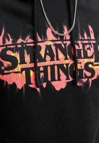Schwarzer Hoodie mit einem "Stranger Things"-Grafikdesign in feurigen Rot- und Orange-Tönen. Enthält Schnürsenkel und eine Kettenakzent.