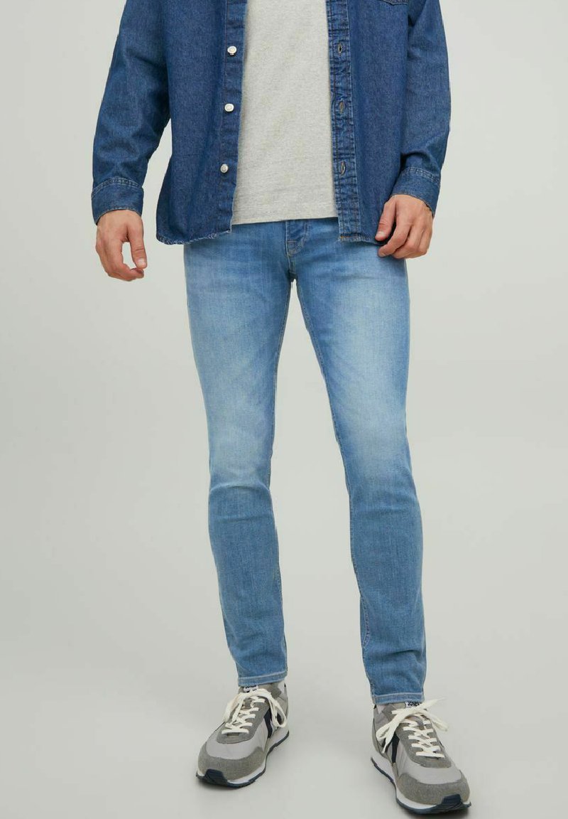 Jeans de mezclilla azul claro con corte slim, combinados con una camiseta gris y una camisa de botones azul oscura, complementados con zapatillas gris y blancas.