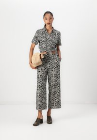 Chemise à manches courtes à motif de losanges en noir et blanc, nouée à la taille, associée à un pantalon à jambes larges ; sac en toile naturel et chaussures texturées.