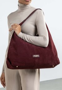 Burgundy suede shoulder bag with a slouchy design, gold hardware accents, and a small logo tag. Soft texture and spacious shape.