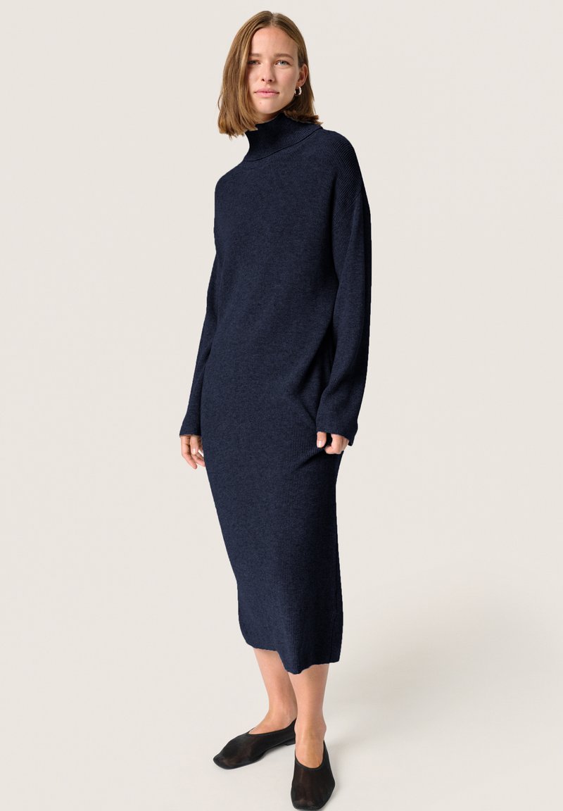 Rochie tricotată bleumarin, cu guler înalt, mâneci lungi și o siluetă dreaptă, asortată cu pantofi slip-on negri.