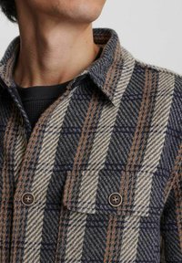 Wool košulja-jakna s uzorkom houndstooth u tamnoplavoj, bež i narančastoj boji. Dugmad na dva džepa na prsima; teksturirani materijal.