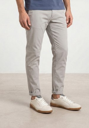 Man draagt lichtgrijze slim-fit broek met opgerolde pijpen en witte sneakers met rubberen zolen, staand op een lichte vloer.