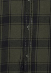 Groene en zwarte geruite flanellen shirt met vijf knopen, zichtbare stiksels en een zachte textuur. Het patroon bestaat uit grote horizontale en verticale lijnen.