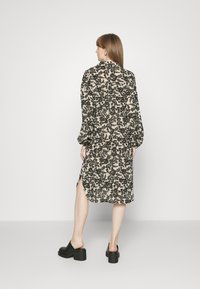 Vestido preto e creme com padrão floral, de mangas longas, fendas laterais e um corte solto. Usado com mules pretos de salto bloco sobre um fundo cinza.