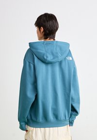 Haut à capuche surdimensionné en teal avec cordon de serrage et logo brodé dans le haut du dos. Fabriqué en mélange de coton, avec des poignets et un ourlet côtelés.