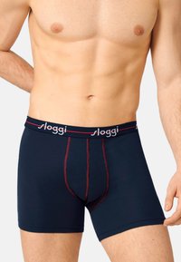 Männlicher Oberkörper, der marineblaue Boxershorts mit roter Naht und einem elastischen Bund trägt, auf dem "sloggi" steht, vor einem schlichten Hintergrund.
