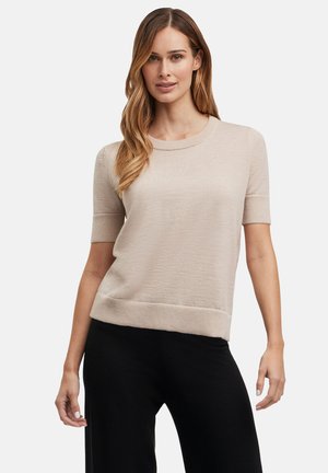 Beżowy sweter z krótkim rękawem i okrągłym dekoltem, wykończony ściągaczem, uszyty z miękkiego, teksturowanego materiału dziewiarskiego. W zestawieniu z czarnymi spodniami.