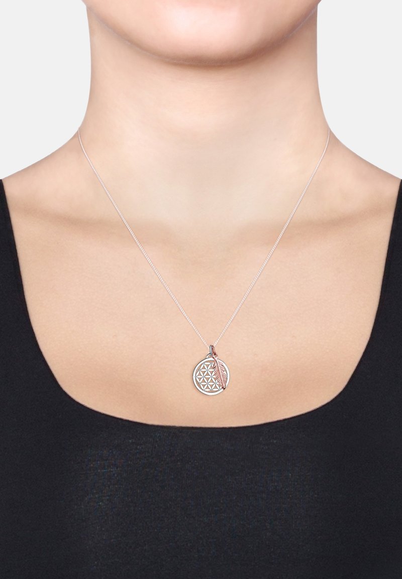 Silberne Halskette mit einem runden Anhänger mit geometrischem Muster und einem Akzent in Roségold in Form einer Feder, getragen auf einem schwarzen Oberteil.