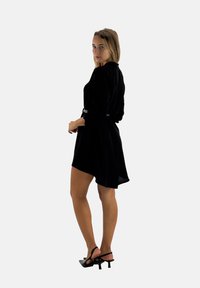 Robe noire à manches longues avec une taille ceinturée et un ourlet asymétrique. Portée avec des talons noirs à lanières. Tissu lisse avec un léger éclat.