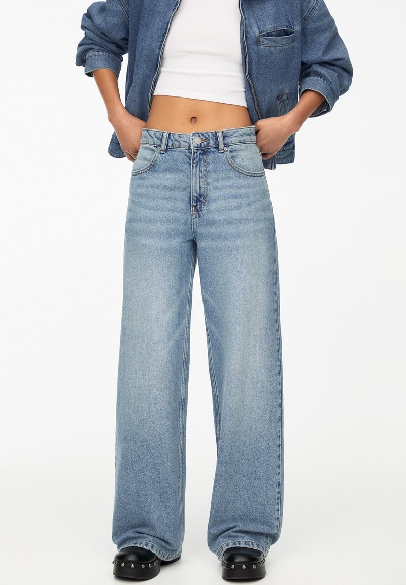 Personne portant un jean bleu clair taille haute à jambes larges, un top court blanc, une veste en denim et des chaussures noires cloutées, les mains dans les poches.