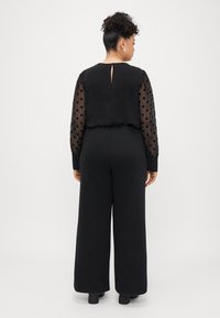 Czarny kombinezon z solidnym topem i dekoltem w kształcie łezki, przezroczystymi długimi rękawami w kropki oraz szerokimi nogawkami. Gładka tekstura w całym ubraniu.