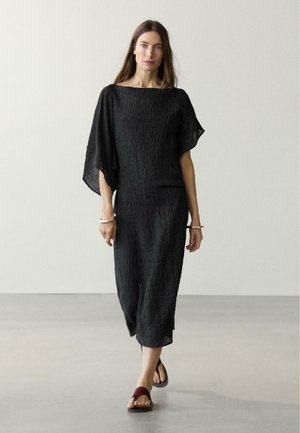 Frau, die ein strukturiertes schwarzes Midikleid mit weiten Ärmeln, silbernen Armbändern und burgunderfarbenen offenen Sandalen vor einem schlichten Hintergrund trägt.