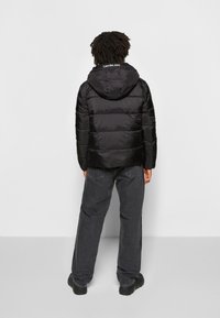 Giacca a piumino nera con cappuccio, caratterizzata da trapuntatura orizzontale e un logo sul colletto. Abbinata a pantaloni di denim scuro larghi e calzature nere.