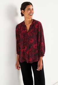 Rød blomsterbluse med V-hals, lange puffede ærmer og løstsiddende pasform, med en blanding af blomster- og prikkede mønstre på en sort baggrund.