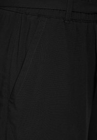 Černé látkové kalhoty s hladkou texturou, které mají boční kapsy a elastický pas. Podrobnosti šití jsou viditelné podél švů.