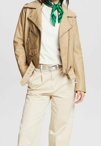 Kobieta w beżowej skórzanej kurtce nosi białą bluzkę, beżowe spodnie w wysokim stanie z paskiem oraz zielony, wzorzysty szalik na szyi. Ręka w kieszeni.