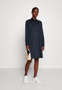 Marineblauwe blousejurk met lange mouwen, een kraag en een rechte silhouette, gecombineerd met witte sneakers en een crèmekleurige handtas.