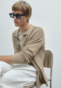 Beige Strickjacke mit strukturiertem Finish, über einem hellbeigen Hemd getragen. Schwarze rechteckige Sonnenbrille. Weiße Hose und ein Metallstuhl.