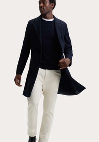 Marinkappa med slitsad krage, enkelknäppt modell, bärs över en mörk tröja och ljusa byxor. Slät tyg, avslappnad passform.