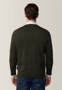 Pull en maille vert avec un col rond et des poignets côtelés, porté par-dessus une chemise col bleu clair. Un jean foncé est également visible.