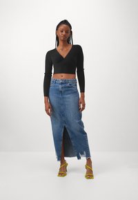 Schwarzes, geripptes Crop-Top mit langen Ärmeln, kombiniert mit einem hellblauen Denim-Maxirock mit einem Schlitz vorne und fransigem Saum. Gelbe Riemchenabsätze.