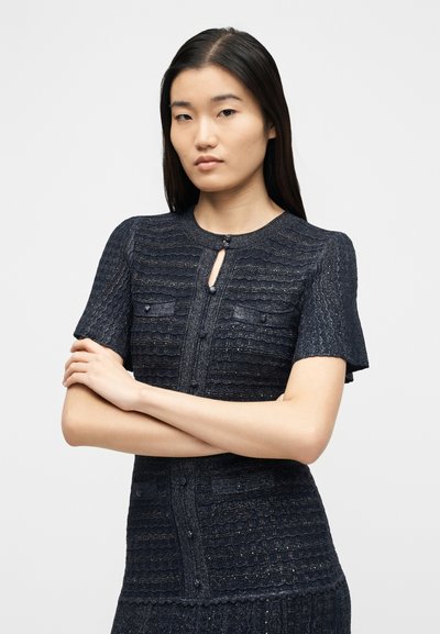 Γυναίκα με μακριά σκουρόχρωμα μαλλιά, που φοράει υφασμάτινο μπλε σκούρο κοντομάνικο φόρεμα με κουμπιά, όρθια με σταυρωμένα τα χέρια μπροστά σε απλό φόντο.