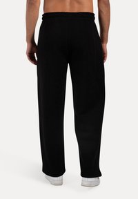 Sorte træningsbukser med elastisk talje. Løs pasform med lige ben. Letvægtsmateriale og minimale syninger. Hvide sneakers synlige.