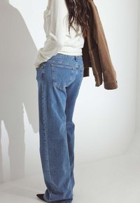 Cilvēks ģērbjas zilās džinsu biksēs un baltā garajās piedurknēs topā, ar brūnu žaketi, kas uzkārta uz viena pleca, pret baltu fona.