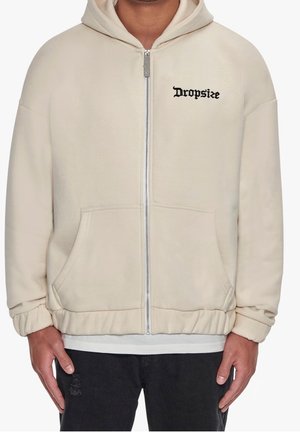 Sweat à capuche beige zippé avec poches avant et texte noir « Dropsize » sur la poitrine gauche, porté sur une chemise blanche et associé à un pantalon noir.