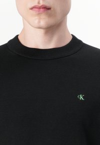 Gros plan d’une personne portant un pull ras-du-cou noir avec un petit logo « cK » vert brodé sur la poitrine.