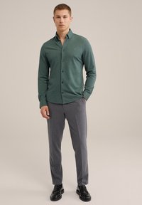 Camisa verde de manga larga con cuello abotonado, botones negros, combinada con pantalones de vestir grises y zapatos negros. Textura simple y diseño ajustado.