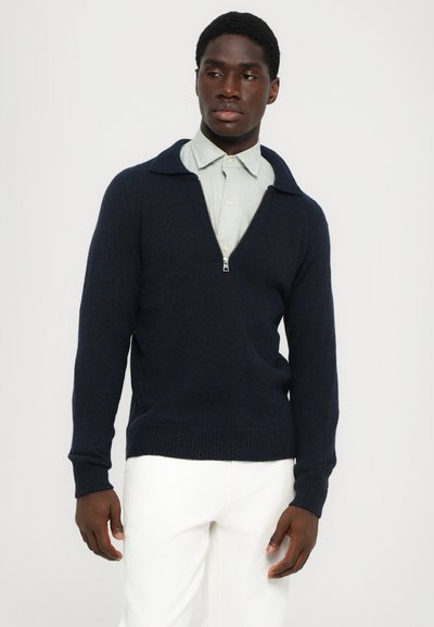 Suéter de lana azul marino con cuello con cremallera, puños acanalados y dobladillo. Lleva puesto una camisa de cuadros en verde claro y se combina con pantalones blancos.
