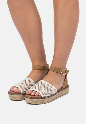 Gambe con sandali con zeppa beige, cinturini anteriori in mesh bianco e cinturini alla caviglia color cuoio con fibbie rotonde dorate.