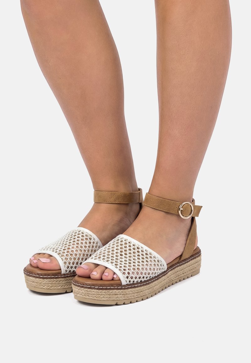 Piernas con sandalias beige de plataforma, con tiras frontales de malla blanca y tiras en el tobillo color canela con hebillas redondas doradas.