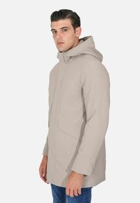 Uomo che indossa una giacca beige chiaro con cappuccio e tasche frontali, e jeans blu, in piedi davanti a uno sfondo bianco uniforme.