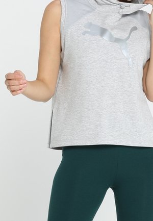 Ærmeløs, lysegrå hættetrøje med metallisk Puma-logo båret sammen med mørkegrønne leggings, viser overkrop og maveområde.