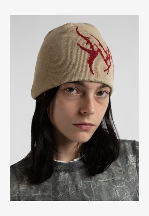 Beżowa dziana czapka z czerwonym abstrakcyjnym wzorem; miękka tekstura i dobrze dopasowany krój. Model ma na sobie przezroczystą, wzorzystą koszulę pod nią.