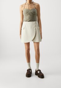 Des chaussures à plateforme marron, un top sans manches olive avec un design froncé, et une jupe mini enveloppante crème avec trois boutons et des poches latérales.