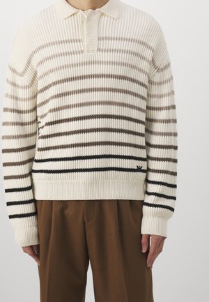Homme portant un pull en maille côtelée crème avec des rayures horizontales marron et noires, associé à un pantalon marron, debout devant un fond uni.