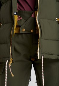 En nära bild av en person som bär en olivgrön puffjacka och matchande byxor, med guldknäppningar och synliga dragsko.